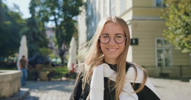 Bonita joven rubia alegre y alegre con gafas mirando a la cámara con una sonrisa sincera en el fondo del antiguo edificio de la ciudad en un día soleado — Vídeos de Stock