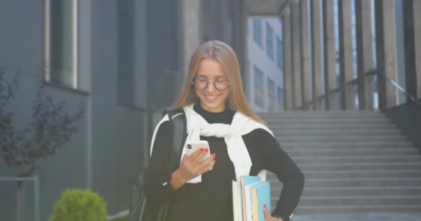 Gros plan d'attrayant sourire heureuse jeune femme aux cheveux clairs moderne dans des lunettes qui tient des livres dans les mains et en revoyant des vidéos drôles sur mobile près de l'immeuble de la ville — Video