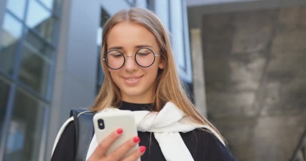Μπροστά όψη του όμορφου χαμόγελου χαρούμενη νέα μοντέρνα ξανθούλα σε γυαλιά που στέκεται κοντά στο σύγχρονο κτίριο της πόλης και χρησιμοποιώντας το smartphone της — Αρχείο Βίντεο