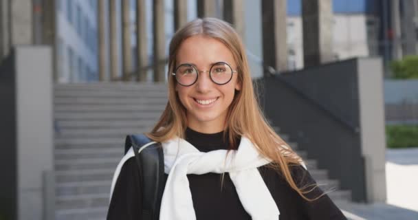 Widok z przodu piękny uśmiechnięty szczęśliwy nowoczesny młoda blondynka w casual ubrania i okulary, które pozowanie przed kamerą na tle biurowca miasta — Wideo stockowe