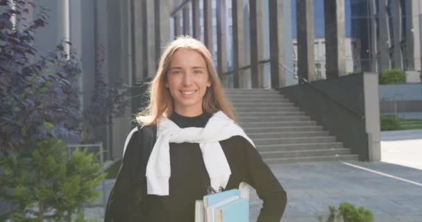 Pretty smiling happy young light-haired girl in casual clothes with books standing in front of city modern building and looking into camera — Stock Video