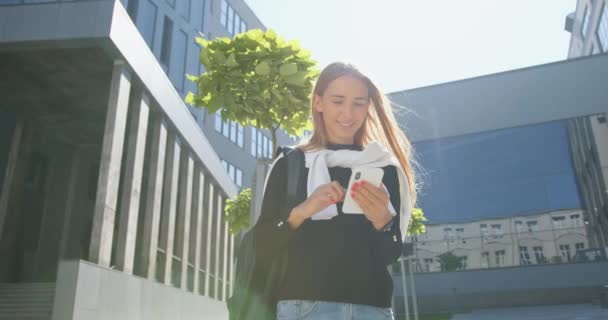 Belle jeune fille souriante et satisfaite aux cheveux clairs en vêtements décontractés avec sac à dos debout près de l'immeuble de bureaux et utilise son mobile le jour ensoleillé — Video