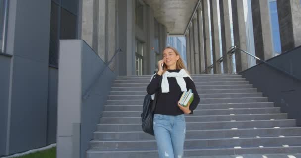 Krásná usmívající se spokojená mladá světlovlasá dívka-student v ležérní oblečení jít dolů po schodech, zatímco opouští univerzitu a těší její telefonní rozhovor — Stock video