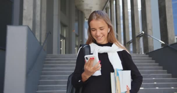 대학이나 사무실 건물 계단을 내려가는 동안가벼운 긴 머리를 하고 책을 가지고 다니고 전화 놀이를 즐기는 멋진 행복 한 소녀 — 비디오