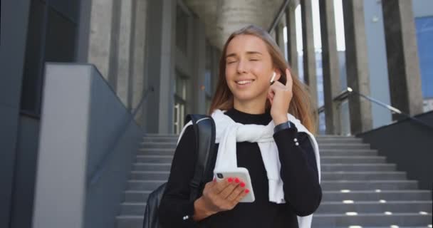 Lovely θετική χαρούμενη κομψή νεαρή ξανθούλα με σακίδιο ακούγοντας μουσική στα ακουστικά, ενώ αφήνοντας σύγχρονη πόλη κτίριο, αργή κίνηση — Αρχείο Βίντεο