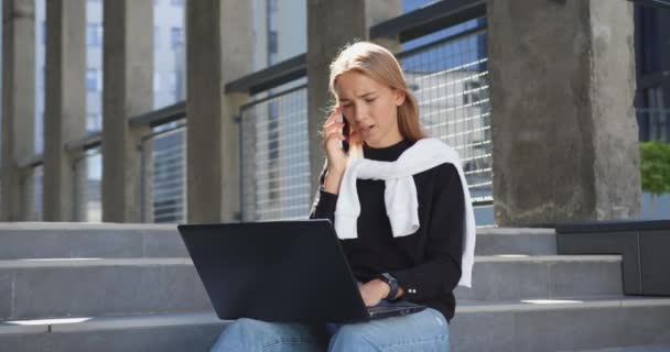 Mooi zelfverzekerd glimlachend positief jong blondje in casual kleding zittend op gebouwen stappen, praten over de telefoon en werken op de computer buiten — Stockvideo