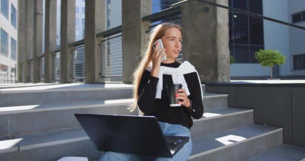 Attraktivt leende nöjd ung ljushårig flicka i trendiga ungdomliga kläder sitter på byggnader steg, arbetar på laptop, dricka kaffe från pappersmugg och prata i telefon — Stockvideo