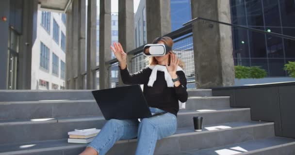 Портрет сучасної розумної молодої жінки в повсякденному одязі, яка сидить на сходах будівель у гарнітурі віртуальної реальності і працює на уявному екрані — стокове відео