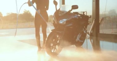 Siyah elbiseli hoş, modaya uygun genç esmer gün batımında yüksek basınçlı su tüpüyle serin siyah motosikletini self-servis istasyonunda yıkıyor.