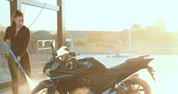 Adorable morena joven positiva satisfecha en ropa negra lavando su costosa moto negra en auto-servicio de lavado de coches con tubo de chorro de agua al atardecer — Vídeos de Stock