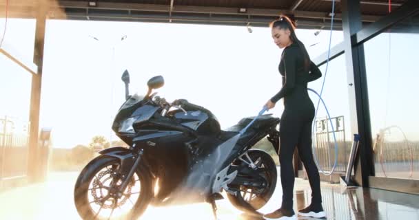 Досить задоволена стильна молода брюнетка з високим хвостом і чорним носінням миття спортивного чорного мотоцикла на автомийці самообслуговування з водопровідною трубкою вдень — стокове відео