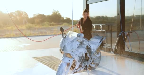 Okouzlující pozitivní bezstarostné mladé brunetky v černém oblečení mytí její sportovní černý motocykl s pěnou na self-service mytí aut ve dne, zblízka — Stock video