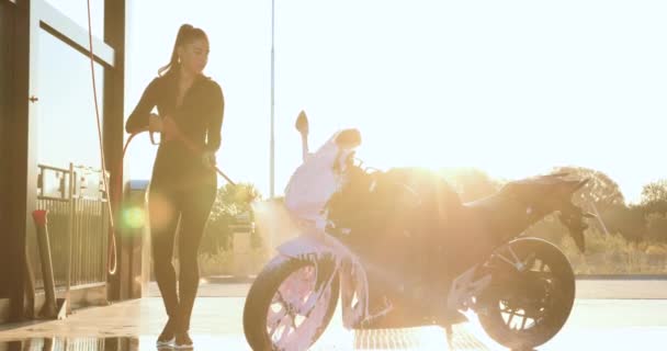 ハイポニーテールの黒い服で魅力的なスタイリッシュな若いブルネットは、日中にセルフサービスの車の洗濯で泡でスポーティーで豪華なバイクを洗う — ストック動画