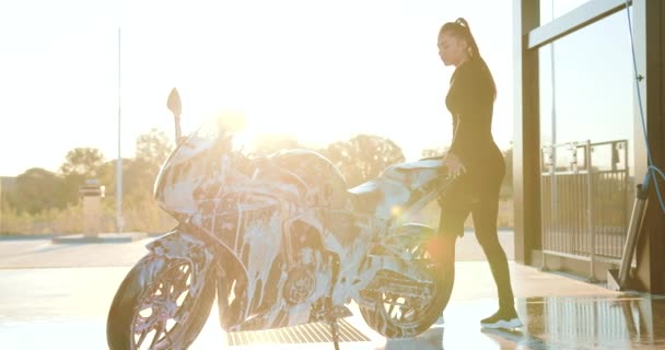 Привлекательная улыбающаяся молодая стильная брюнетка в черной одежде, стоящая рядом со своим крутым спортивным мотоциклом, покрытым пеной, пока стирает его на автомойке самообслуживания. — стоковое видео