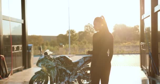 Krásná usměvavá mladá brunetka v černém oblečení pózuje v blízkosti svého drahého černého železného koně pokrytého pěnou při mytí na myčce aut při západu slunce — Stock video