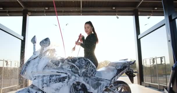 Samoobslužné mytí aut venku koncept, kde nádherné štíhlé stylové mladé brunetky v černém oblečení mytí její sportovní černá motorka s pěnou — Stock video