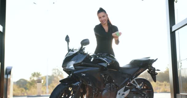Портрет красивой сексуальной улыбающейся брюнетки 25 лет в черной одежде, которая вытирает тканью свой современный черный мотоцикл на автомойке самообслуживания на закате — стоковое видео