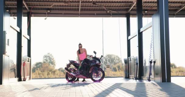 Blick aus der Ferne, wo schöne verführerische schlanke 25-jährige Mädchen in Sonnenbrille und rosa Top Waschen luxuriösen sportlichen Motorrad mit schäumender Lösung auf Autowäsche im Freien — Stockvideo
