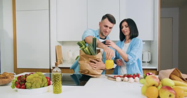 Atractiva feliz sonriente pareja casada desempacar bolsa de papel de comida llena de diferentes verduras en la cocina contemporánea cuando llegó a casa del mercado — Vídeos de Stock