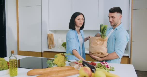 Lovely happy smiling brunette helping to unpack food paper bags her positive husband which came back from market and they together standing near dinner table in the kitchen — Stock Video