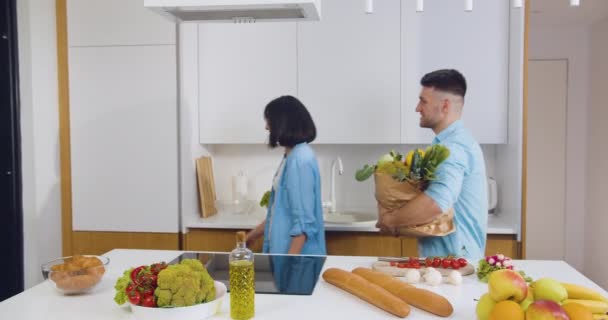 Encantadora pareja sonriente feliz en el amor regresó del mercado de alimentos con una gran bolsa de papel llena de alimentos frescos y poner todos los productos en la nevera — Vídeos de Stock