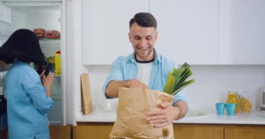 Güzel görünüşlü, mutlu, mutlu, evli bir çift yemek paketini açarken ve taze sebzeleri buzdolabına koyarken birlikte eğleniyorlar.