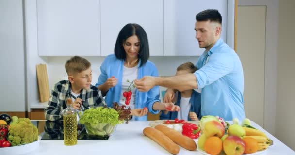 Bonito alegre dois meninos ajudando sua mãe e pai a cozinhar salada de legumes para o jantar de família na cozinha contemporânea, conceito de família, vista frontal — Vídeo de Stock