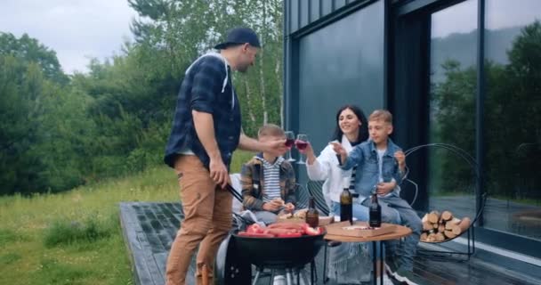 Idilio familiar donde feliz padre sonriente preparando verduras y salchichas en la parrilla y tintineo vasos con vino con su esposa, mientras que dos hijos comiendo pizza al aire libre — Vídeo de stock