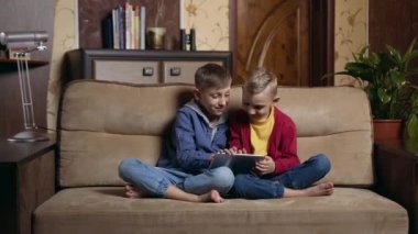 Attractive smiling small two boys sitting on soft couch at home and use tablet pc to play online games,leisure concept