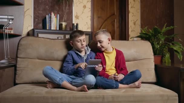 Pleasant satisfied small two boys in casual clothes resting on soft couch in contemporary room and using tablet device to watch cartoon or play video game — Stock video