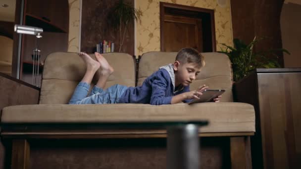 Attractive happy smiling 12-aged smart boy lying on the couch at home and watching cartoon or funny video on tablet pc ,front view — Vídeo de Stock
