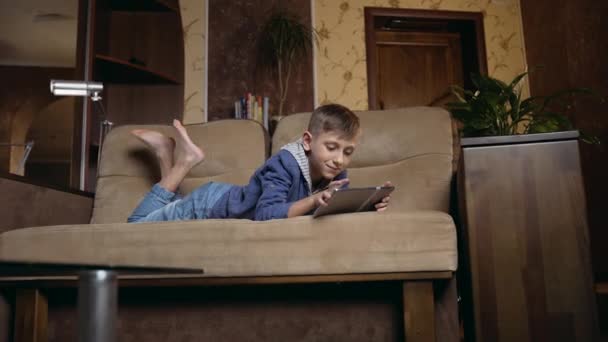 Good-looking positive carefree 12-aged boy in casual clothes relaxing on soft couch at home and uses tablet computer ,close up — 图库视频影像