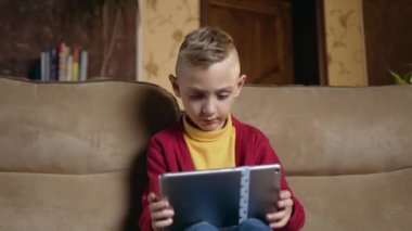 Portrait of handsome concentrated small boy in homewear which sitting on comfortable couch at home and uses tablet device,close up