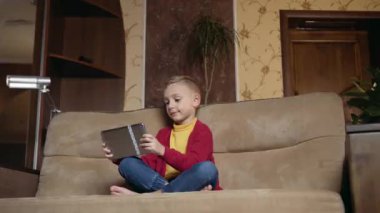 Attractive positive small boy relaxing on comfortable sofa in contemporary living-room and playing online video game on tablet pc,close up