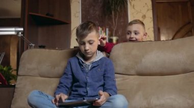 Attractive calm modern 12-aged boy sitting on the couch at home and uses tablet pc to watch video or gaming,another boy interfering him and grabbing by ears