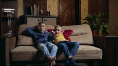 Front view of attractive positive calm two brothers which relaxing on soft couch at home and watching TV,one of boys presses the remote control from TV set