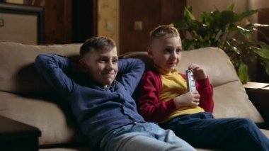 Çekici, olumlu, gülümseyen iki arkadaş ya da kardeşin yan görünüşü. Evdeki yumuşak kanepede dinleniyorlar ve TV programlarını gözden geçiriyorlar.