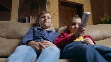Portrait of good-tempered happy smiling schoolboys which watching funny movie or cartoon on TV,one of them changing channel or giving volume using remote control