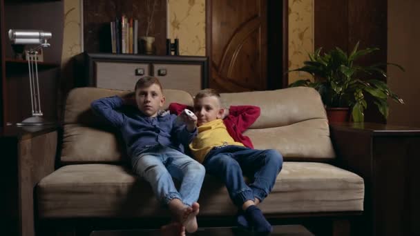 Front view of attractive positive calm two brothers which relaxing on soft couch at home and watching TV,one of boys presses the remote control from TV set — Stock video