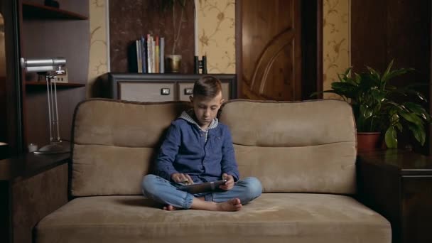 Привлекательный серьезный подросток сидит в позе лотоса на диване дома и использует планшетный ПК, в то время как его младший брат бежит к нему и смотреть, что он делает — стоковое видео