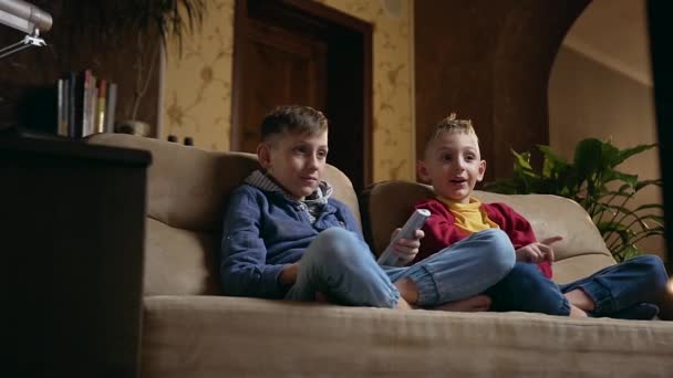 Schön lächelnd entspannen sich zwei Jungs in Hauskleidung zu Hause auf dem Sofa vor dem Fernseher bei einem interessanten Programm oder Film, während sie abends gemeinsame Freizeit verbringen. — Stockvideo