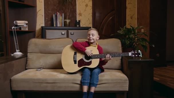 Симпатичный улыбающийся маленький мальчик в домашнем платье сидит дома на мягком диване и играет на гитаре, вид спереди — стоковое видео