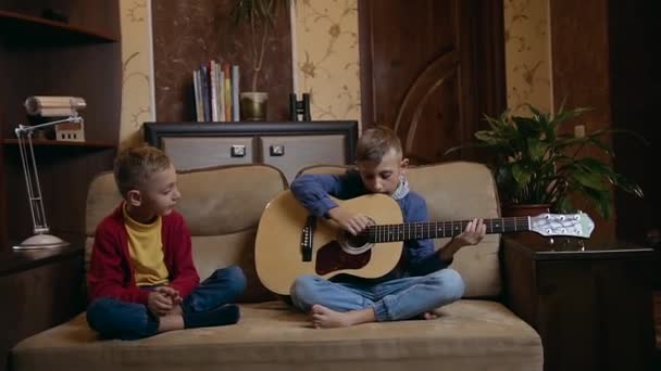 Bello sorridente ragazzo di 10 anni seduto sul divano insieme al fratello maggiore che impara a suonare la chitarra, da vicino — Video Stock