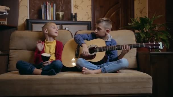 Knappe glimlachende ijverige schooljongens-broers zitten op de zachte bank en spelen op muziekinstrumenten, het doorbrengen van gezamenlijke avond thuis — Stockvideo