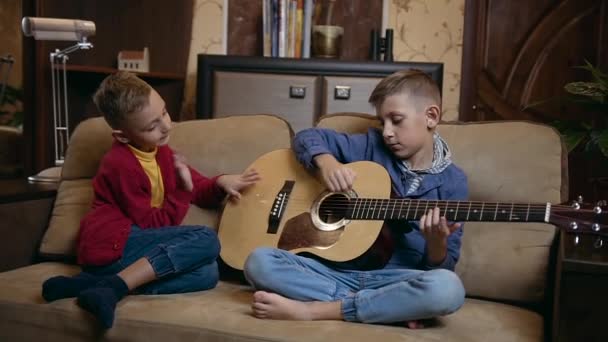 Piękny szczęśliwy nowoczesny dwóch braci w homewears spoczywa na miękkiej kanapie w domu, podczas gdy jeden z nich uczy się grać na gitarze struny, inny pukanie na drewnianej stronie gitary — Wideo stockowe