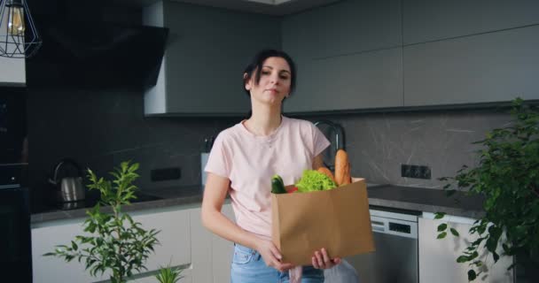 Mooie glimlachende vrolijke vrouw in casual kleding met voedselzak in handen staande in de moderne keuken toen kwam terug van supermarkt — Stockvideo