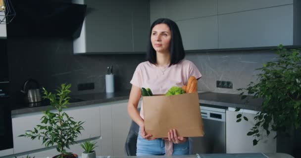 Στοχαστικό όμορφη νεαρή μελαχρινή γυναίκα ποζάρουν στην κάμερα στην κουζίνα με την τσάντα τροφίμων χαρτί που αγοράστηκε στο σούπερ μάρκετ, κοντά — Αρχείο Βίντεο