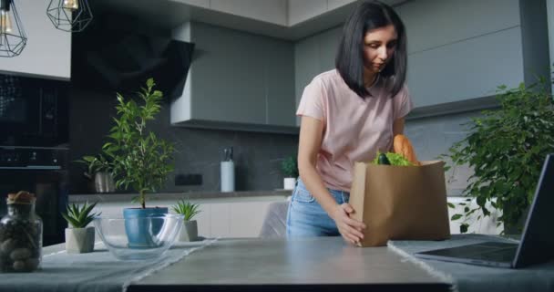 Vacker nöjd ung modern kvinna kom tillbaka från matmarknaden med paket med produkter och packa upp den nära köksbordet — Stockvideo