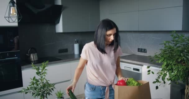 Front view of good-looking confident young brunette which unpacking food bag and putting groceries on kitchen table in glass bowl — Stock Video