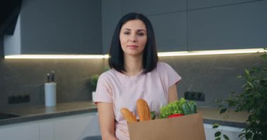 Sevimli, neşeli, siyah saçlı modern kadın kameranın önünde duruyor. Mutfakta ve elinde yiyecek paketi var. Yaklaş.